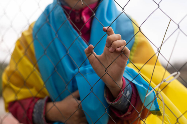triest klein meisje met de vlag van Oekraïne achter een metalen hek. Sociaal probleem van vluchtelingen en gedwongen migranten - Foto, afbeelding