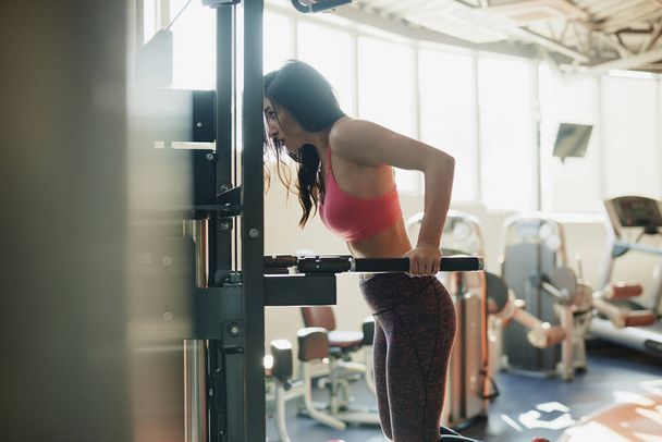 Een sterke vrouwelijke bodybuilder doet pull-ups op een machine in een sportschool. - Foto, afbeelding