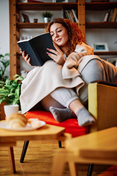 Eine junge Frau mit roten lockigen Haaren, die in die Decke gehüllt ist, liest ein Buch, während sie in ihrem Wohnzimmer auf einem Stuhl sitzt. - Foto, Bild
