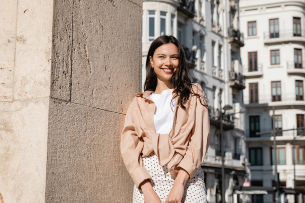 happy woman in beige shirt standing on city street near wall - Foto, afbeelding