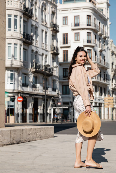 full length of happy woman in trendy clothes holding straw hat on urban street - 写真・画像