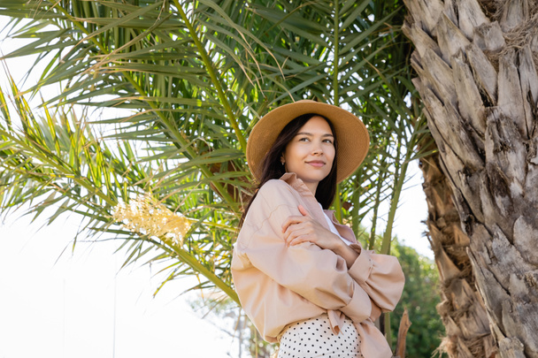 low angle view of woman in straw and beige shirt smiling under palm tree - Fotografie, Obrázek