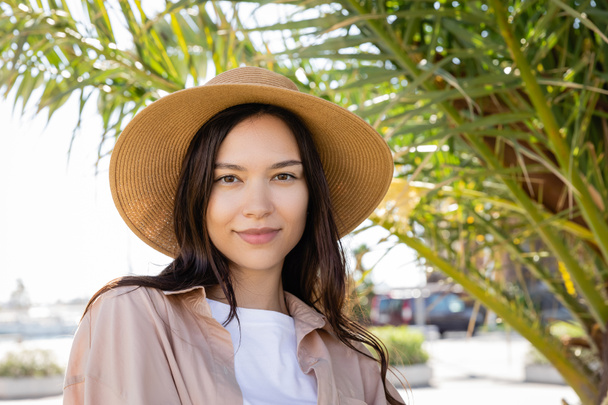 portrait de femme brune positive en chapeau de paille regardant la caméra près de feuilles de palmier floues - Photo, image