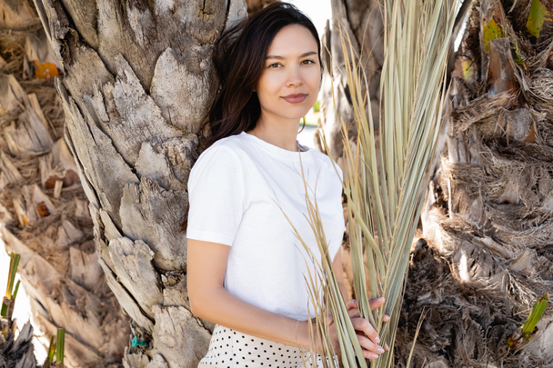 brunette woman in white t-shirt smiling at camera near palm tree trunks - Photo, Image