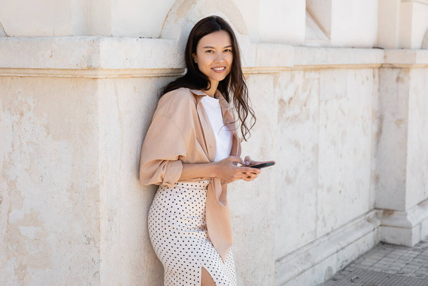 happy and stylish woman standing with smartphone near white wall and looking at camera - Foto, immagini