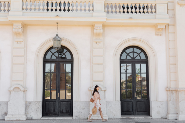side view of woman with handbag and straw hat walking along white building with arch doors - Foto, Imagen