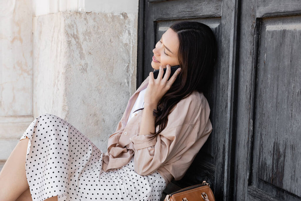 Lächelnde Frau mit geschlossenen Augen sitzt neben schwarzer Holztür und spricht auf Smartphone - Foto, Bild