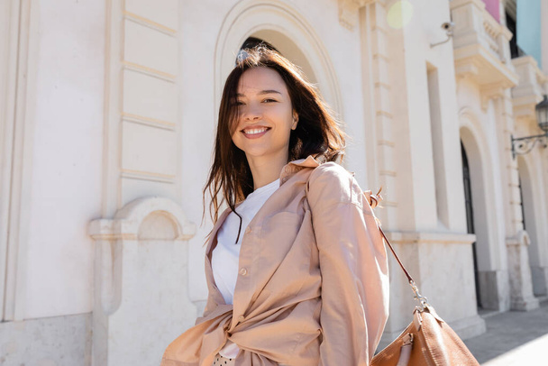 pleased brunette woman in beige shirt smiling at camera near blurred building - Foto, afbeelding