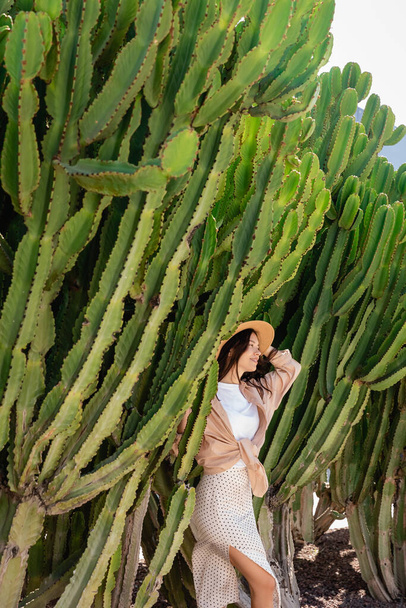 morena mujer en sombrero de paja sonriendo cerca de cactus gigantes en el parque - Foto, imagen