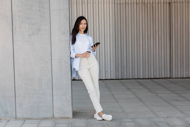 full length of brunette woman standing with smartphone and coffee to go near grey wall - Foto, Bild