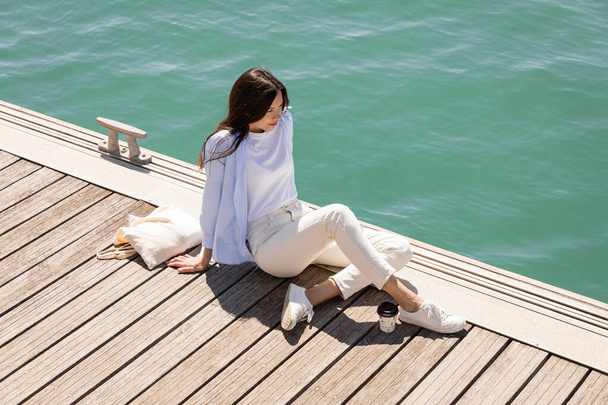 high angle view of woman sitting on pier near paper cup and canvas bag - Photo, Image