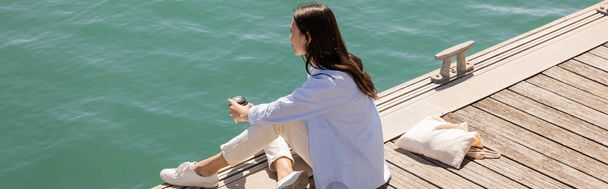 брюнетка жінка дивиться на воду, сидячи на пірсі з виносним напоєм, банер
 - Фото, зображення