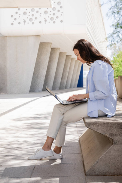 πλαϊνή άποψη της γυναίκας σε λευκό παντελόνι και μπλε πουκάμισο κάθεται με φορητό υπολογιστή στο δρόμο στην πόλη - Φωτογραφία, εικόνα