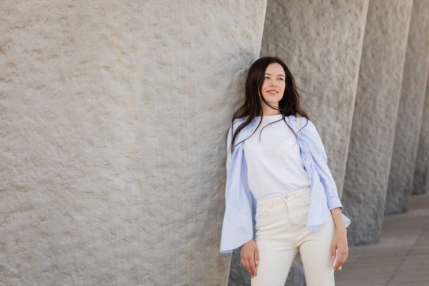 smiling brunette woman in blue shirt and white pants leaning on grey wall - Foto, Bild