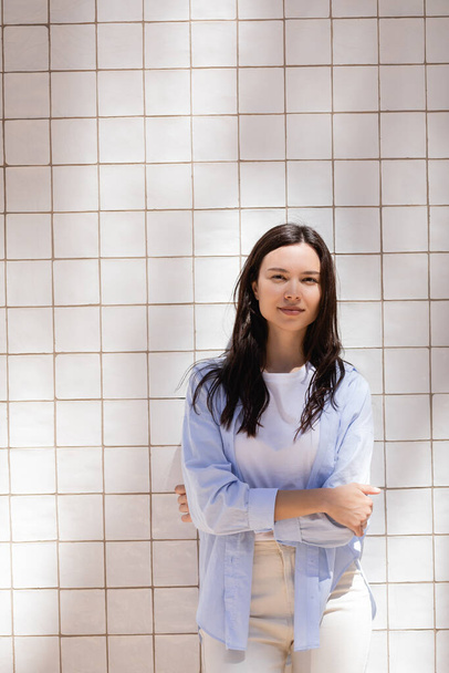 brunette woman in blue shirt standing with crossed arms near white tiled wall - Foto, Imagem