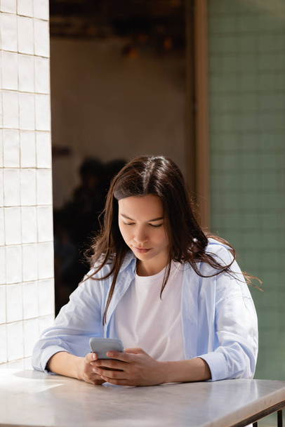 brunette woman in blue shirt messaging on smartphone on cafe terrace - Fotoğraf, Görsel