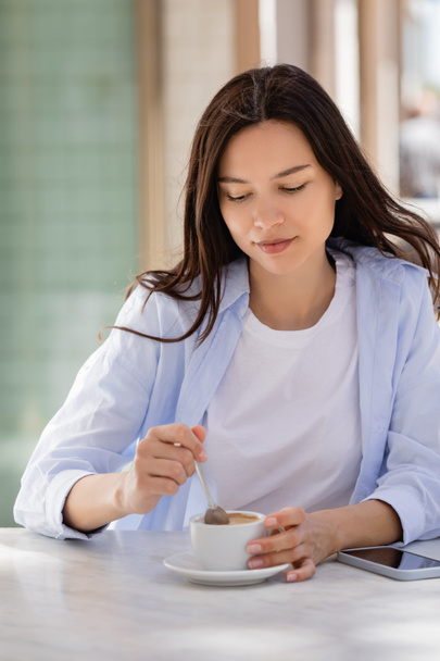 brunette woman holding teaspoon near coffee cup and smartphone with blank screen on cafe terrace - Photo, Image