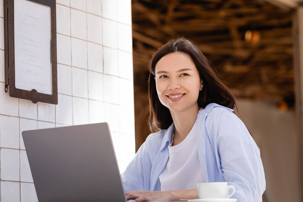 cheerful woman smiling near laptop and coffee cup in street cafe on summer terrace - Foto, Imagem