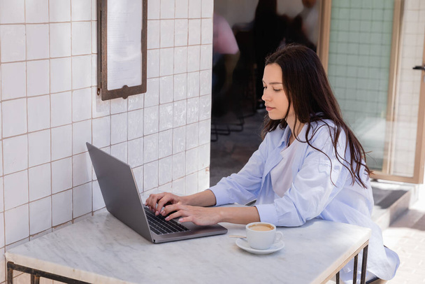 Frau in blauem Hemd tippt auf Laptop neben Kaffeetasse in Straßencafé - Foto, Bild
