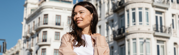 positive woman looking away near white blurred buildings, banner - Photo, Image
