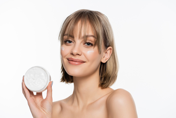 smiling young woman with bare shoulders and cream on face holding container while looking at camera isolated on white - Fotoğraf, Görsel