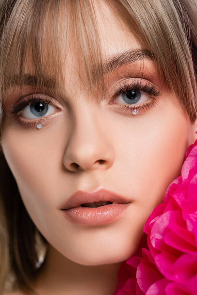 close up of pretty young woman with bangs and shiny rhinestones under blue eyes near pink flower isolated on white - Φωτογραφία, εικόνα