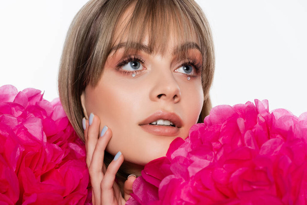 young woman with rhinestones under blue eyes looking away near pink flowers isolated on white - Foto, immagini