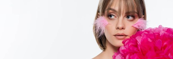 young woman with decorative elements in makeup and pink feathers on cheeks near bright flower isolated on white, banner - Foto, Imagen