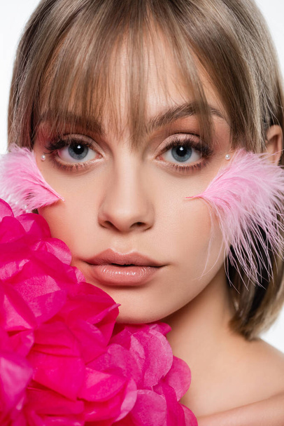 close up of young woman with decorative elements in makeup and feathers on cheeks near pink flower isolated on white - Foto, Imagen
