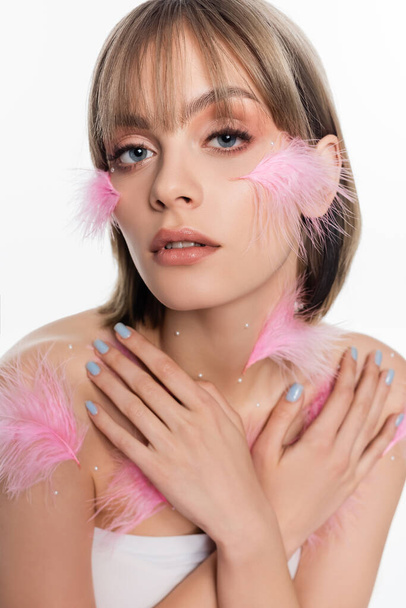 young woman with decorative elements and pink feathers on face and body posing with crossed arms isolated on white - Foto, Imagem