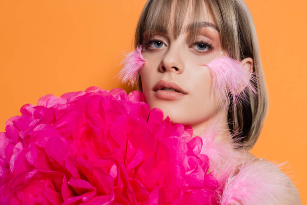 pretty young woman with decorative beads in makeup and feathers on cheeks near pink flower isolated on orange - Foto, Imagen
