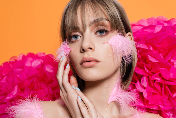 blonde young woman with decorative elements in makeup and pink feathers on cheeks near flowers isolated on orange - Foto, Imagen