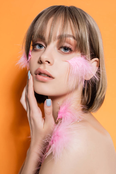young woman with decorative beads in makeup and pink feathers on face isolated on orange - Foto, Imagen