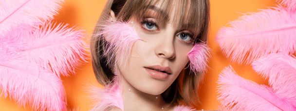 young woman with decorative beads in makeup near pink feathers isolated on orange, banner - Foto, Imagen