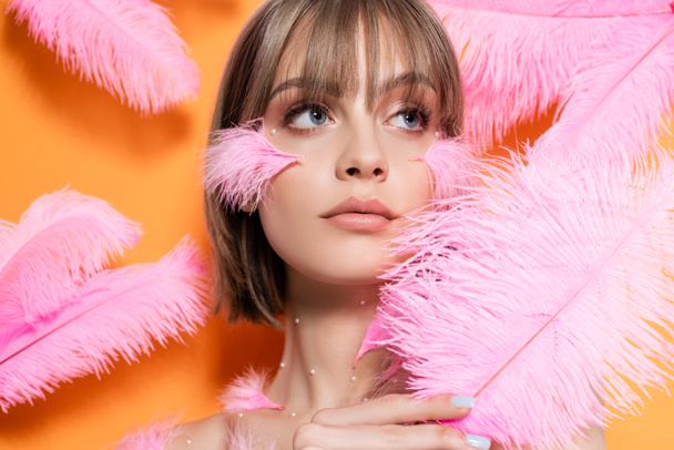 young woman with decorative beads in makeup looking away near pink feathers isolated on orange - Foto, afbeelding