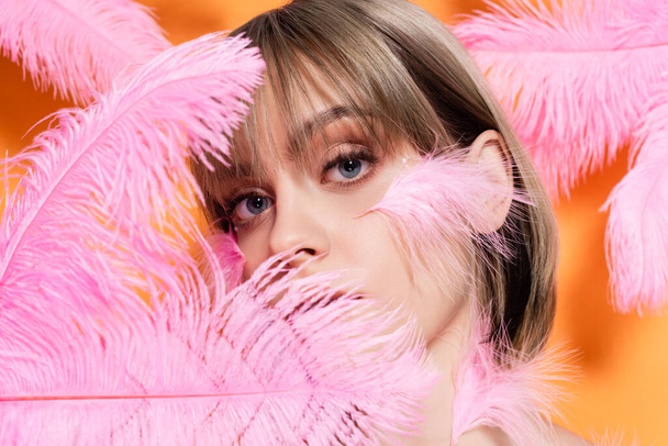 young woman with decorative beads in makeup looking at camera through pink feathers isolated on orange - Foto, Imagen