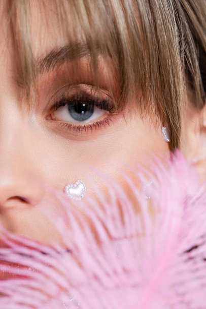 cropped view of young woman with nacreous heart shape elements on cheek near pink feather - Foto, Imagen