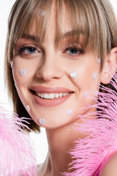 close up of happy woman with nacreous heart shape elements on cheeks near pink feathers isolated on white  - Zdjęcie, obraz