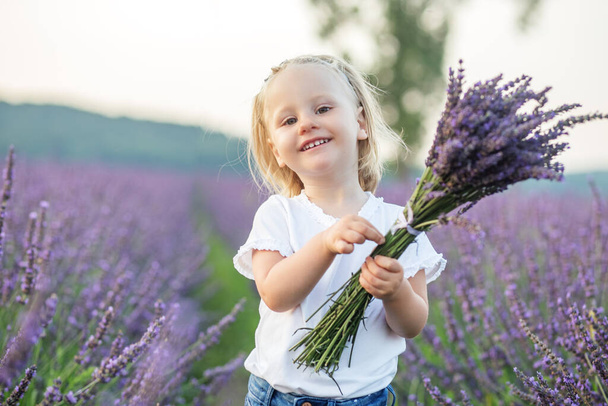 Little girl is walking in lavender field. Children's fantasy. Smiling kid is holding fragrant bouquet of lavender. Close-up portrait of beautiful joyful blond toddler girl. Cheerful child. - Foto, imagen