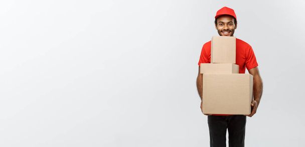 Концепція доставки - Портрет щасливого афроамериканця з доставки в червону тканину, що тримає коробковий пакет. Ізольовано на сірому фоні студії. Копіювати пробіл
. - Фото, зображення