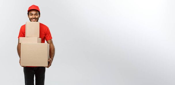 Concepto de entrega - Retrato del hombre de entrega afroamericano feliz en tela roja sosteniendo un paquete de caja. Aislado en Grey studio Background. Copiar espacio
. - Foto, Imagen