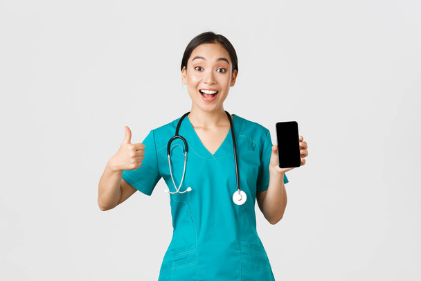 Covid-19, zdravotničtí pracovníci a koncept on-line medicíny. Ohromující a potěšený asijský lékař zkouší nové metody konzultace na dálku během coronavirus, ukazuje smartphone app a palce-up - Fotografie, Obrázek