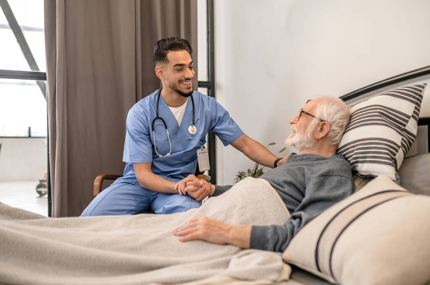 Cheerful medical worker comforting an old man lying in bed - Foto, Imagem