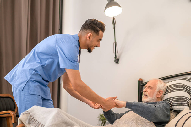 Smiling medical worker lifting the old man from the bed - Photo, image