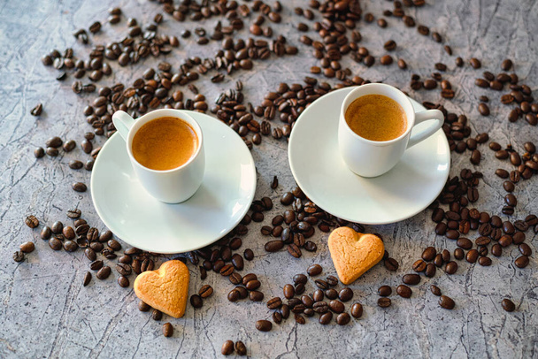 Twee espresso kopjes met schotels op een stenen tafel met hartkoekjes en gebrande koffiebonen. - Foto, afbeelding