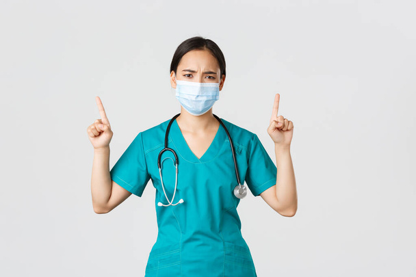 Covid-19, enfermedad coronavirus, concepto de trabajadores de la salud. sombrío decepcionado asiático enfermera, médico en médico máscara y scrubs, señalando los dedos hacia arriba y frunciendo el ceño perplejo, mirada frustrado - Foto, imagen