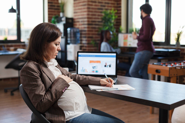 Έγκυος γυναίκα που αναλύει την οικονομική ανάπτυξη της εταιρείας και εξετάζει το οικονομικό και διοικητικό διάγραμμα. - Φωτογραφία, εικόνα
