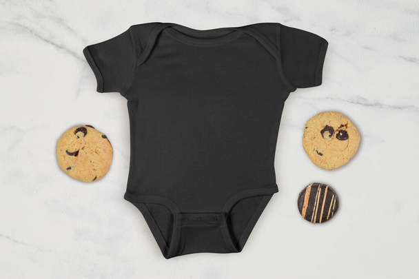 Schwarzer Baby-Onesie umgeben von verführerischem Schokoladenchips und Schokolade gefrosteten Keksen. - Foto, Bild