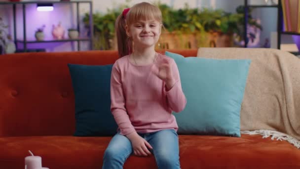 Dziecko dziewczyna siedzi na kanapie w domu patrząc na aparat uśmiechnięty machając rękami gestykulując cześć lub pożegnanie - Materiał filmowy, wideo