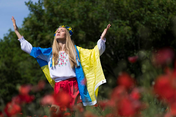 Eine junge blonde Ukrainerin steht auf einem Feld mit roten Mohnblumen und hält die Fahne der Ukraine in der Hand, die ihre Unterstützung für den Krieg in ihrem Heimatland Ukraine zeigt. - Foto, Bild